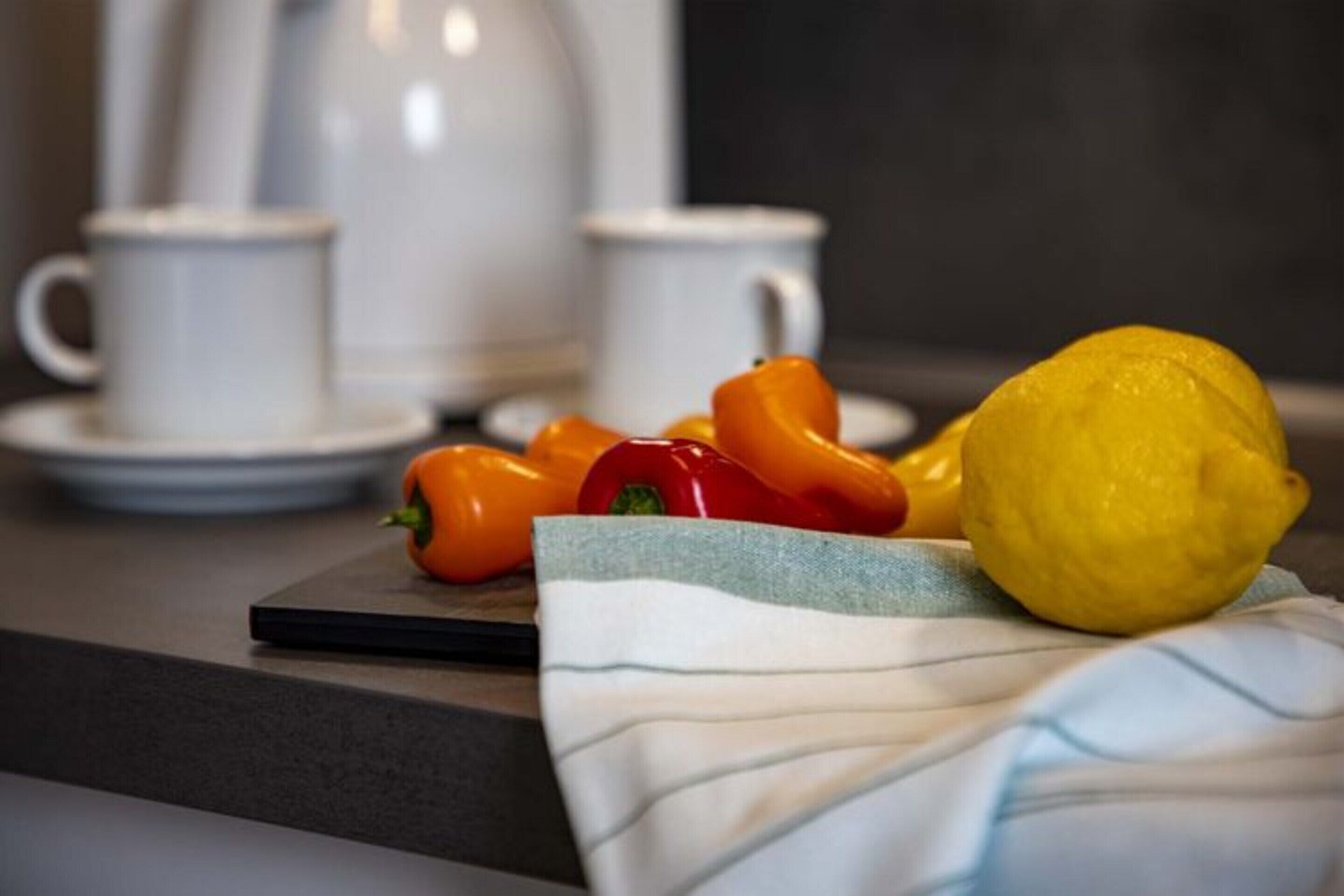 A kitchen counter with a lemon, red and yellow bell peppers, and a cloth. There are two white cups and saucers in the background.