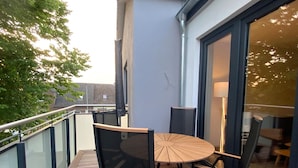 A balcony with a round table and chairs, a glass railing, and a view of the cityscape. - Exclusives Appartement auf Fehmarn (Fehmarn)
