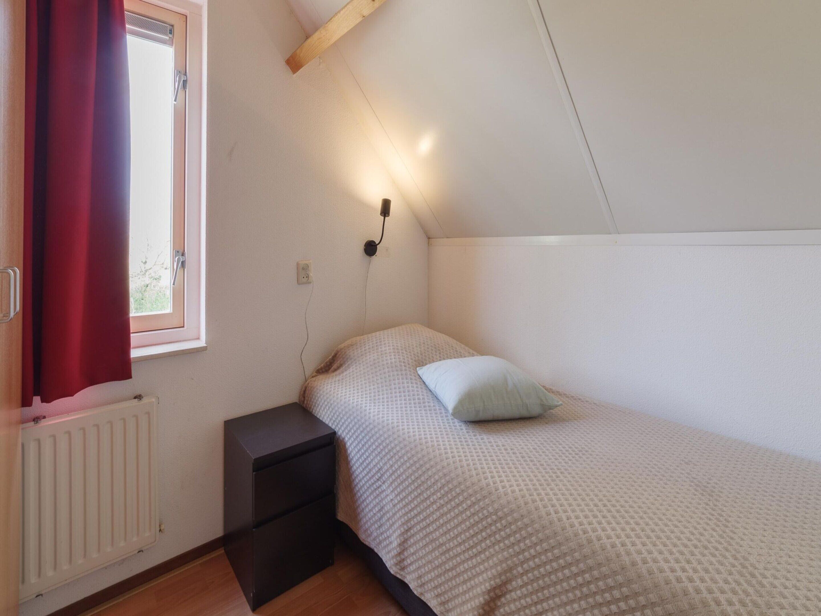 Ein kleines Zimmer mit einem Einzelbett, einem Holz-Kopfteil, einem Nachttisch, einem roten Vorhang und einem Fenster mit Blick auf Gr ünflächen.
