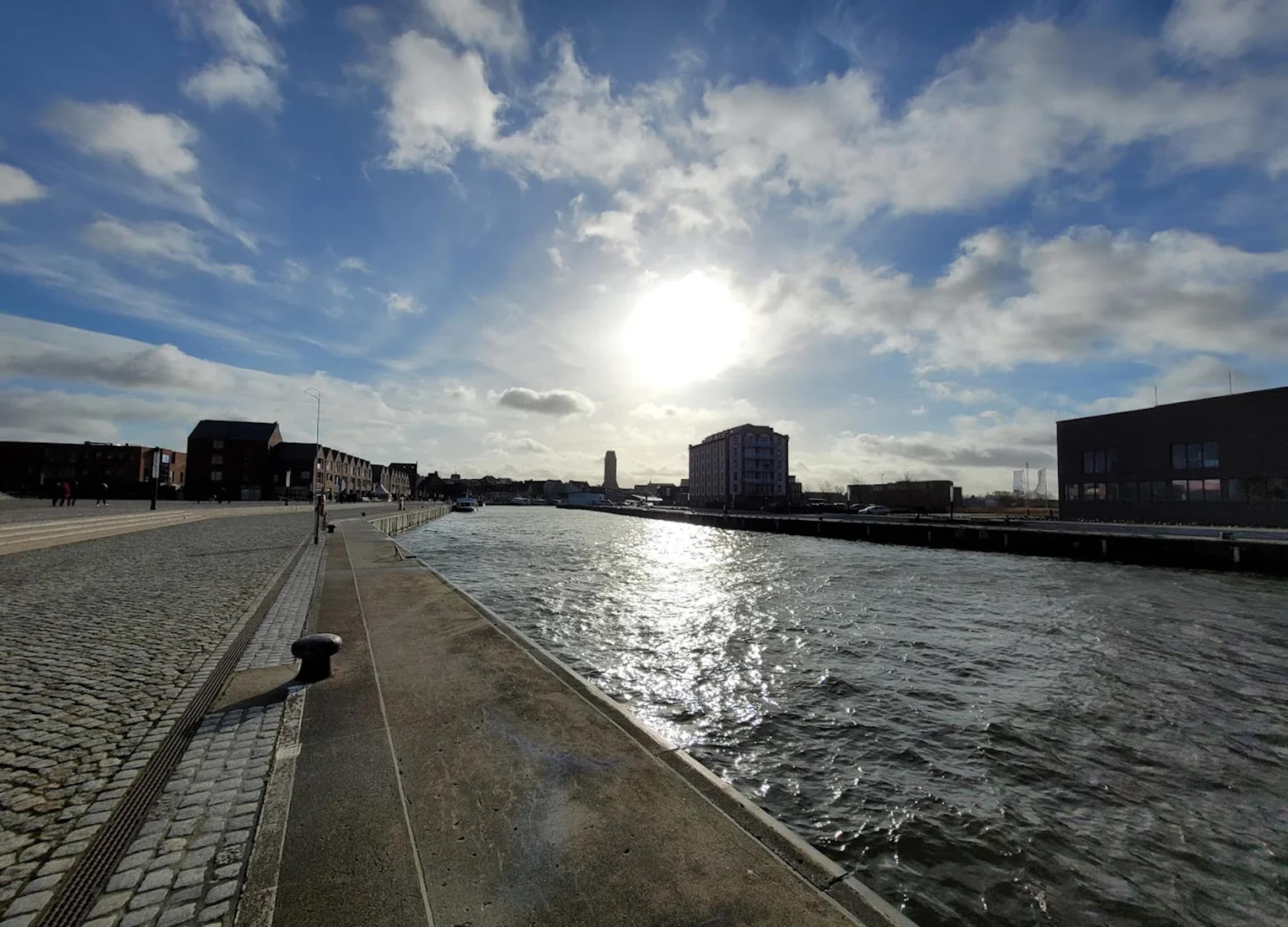 A waterfront promenade with a cobblestone path, a canal, and buildings in the background.
