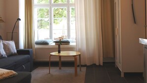 A cozy living room with a sofa, a small round table, a radiator, a window with curtains, and a television mounted on the wall. - Studio in Wismar am Alten Hafen (Wismar)