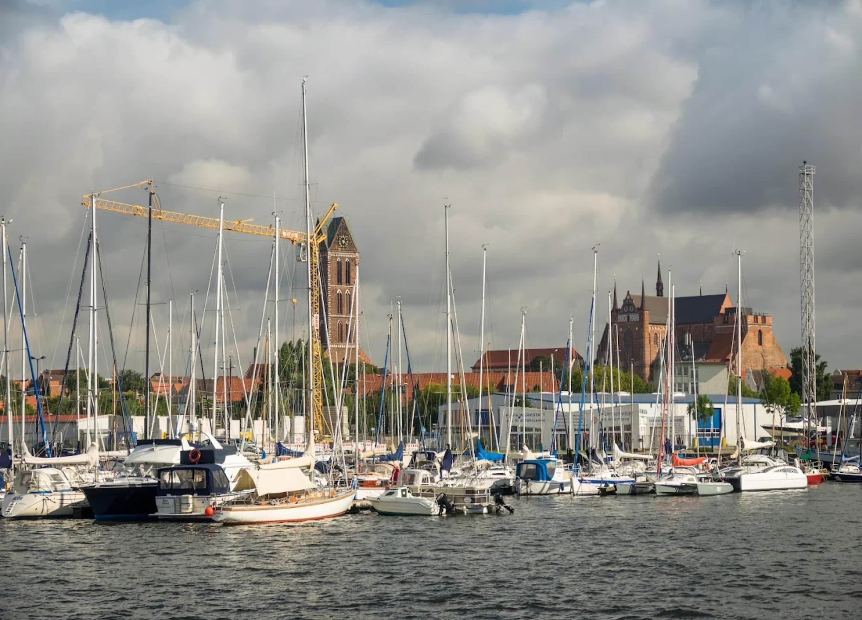 A marina with multiple sailboats docked, a prominent church with a tall bell tower, and a crane in the background.