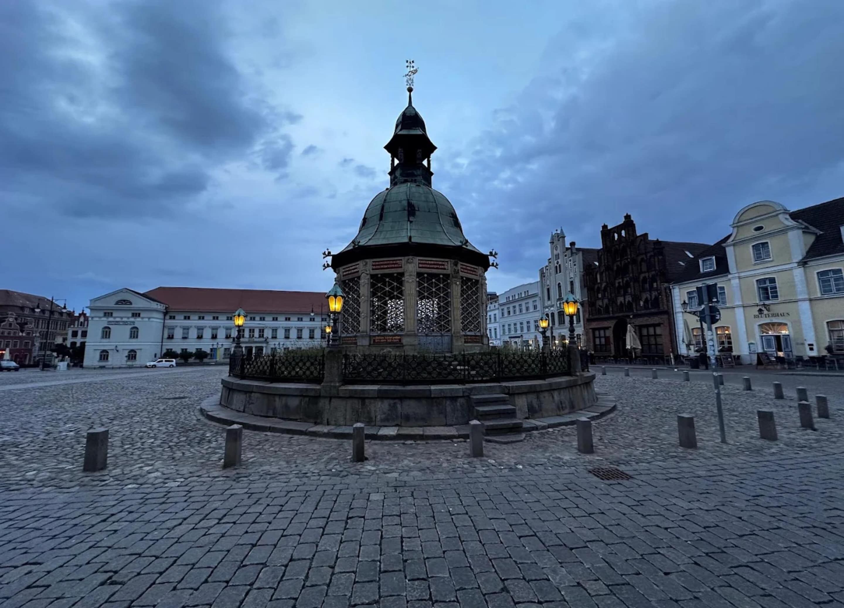 A historical square with cobblestone pavement, a central gazebo with a dome and lanterns, and surrounding classical architecture.