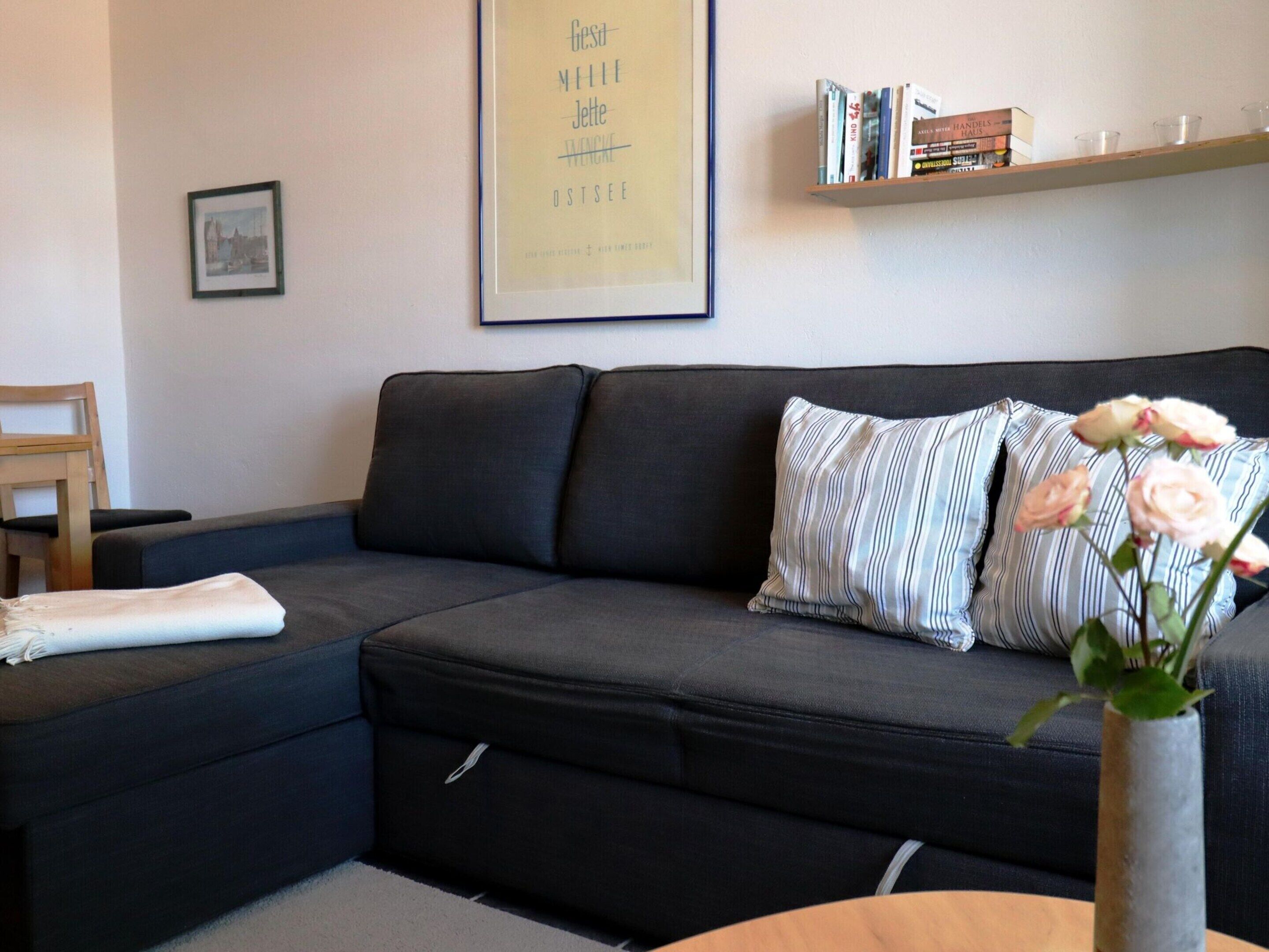 A modern living room with a dark grey sofa, striped pillows, a small table with a vase of flowers, a framed picture on the wall, and a bookshelf with books and decorative items.