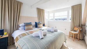 A spacious bedroom with a large bed, a wooden chair, and a view of the sea through a window. - Birdbank Cottage - Two Bedroom Cottage, Sleeps 4 (St Ives)