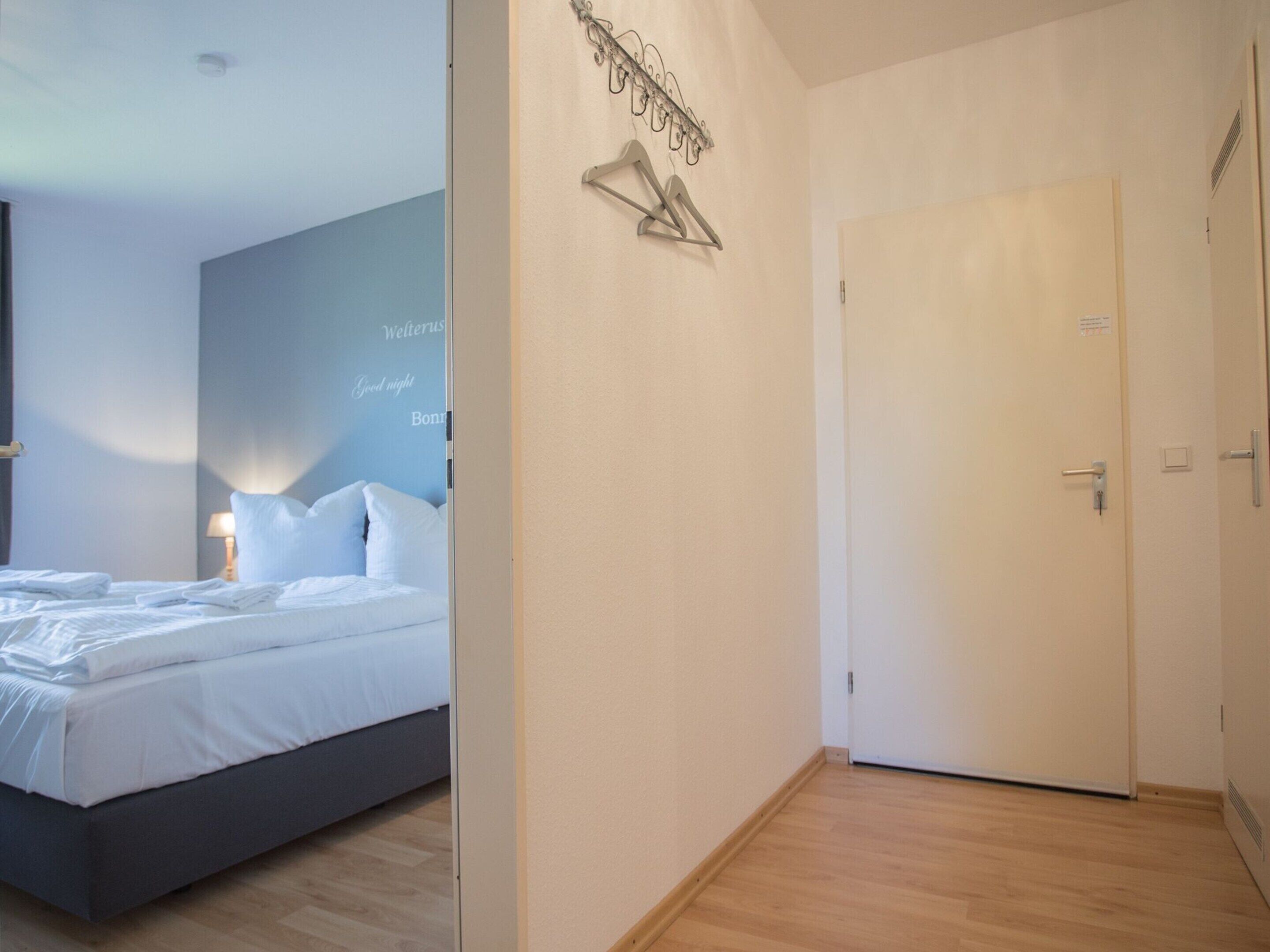 A hotel room with a bed, a bedside table with a lamp, a wall-mounted coat rack, and a door.