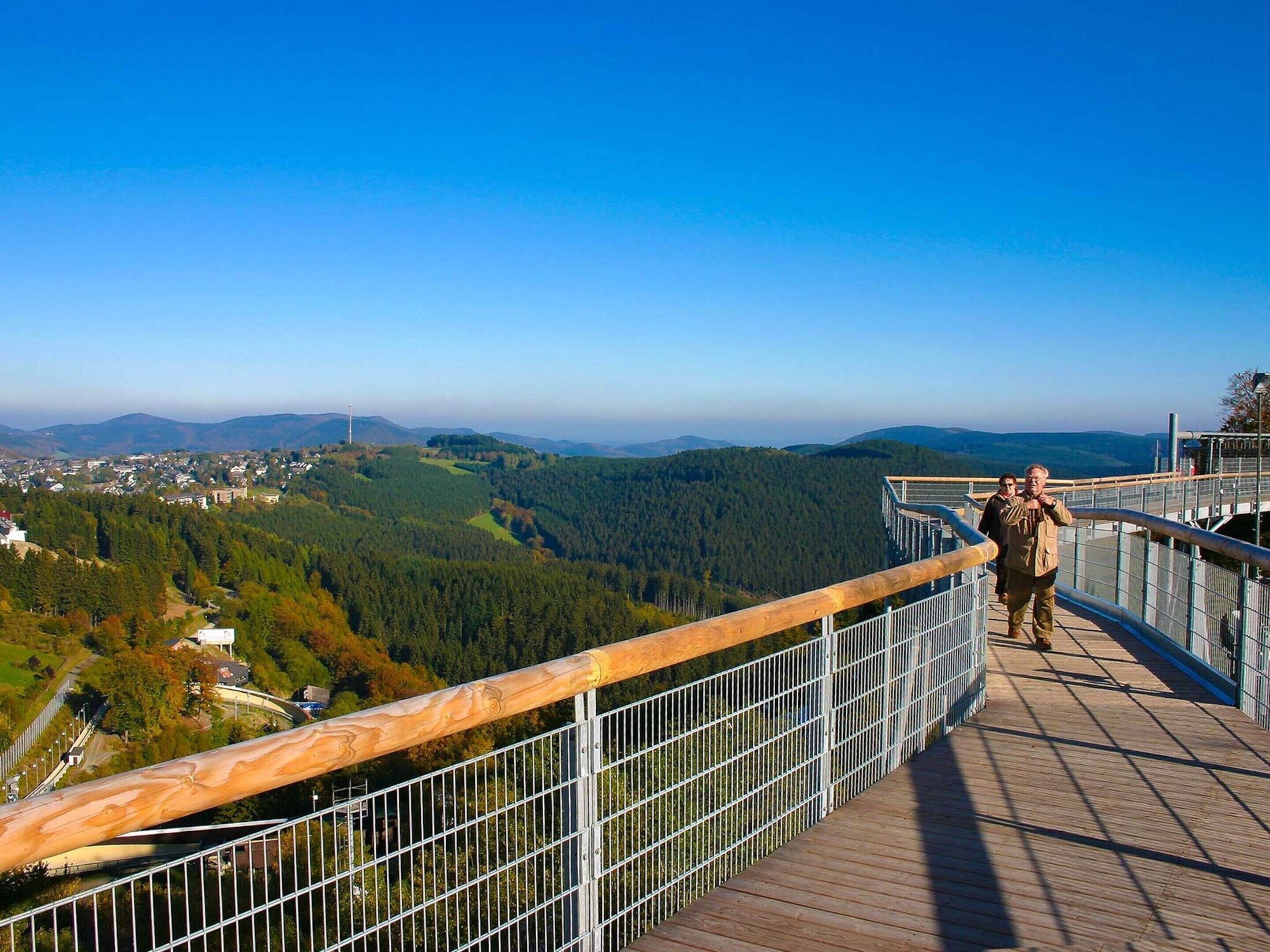 A wooden walkway with metal railings overlooking a forested valley and distant town.