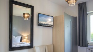 Une chambre d’hôtel avec un lit, une chaise, un grand miroir, un tableau accroché au mur et une fenêtre avec des rideaux.
