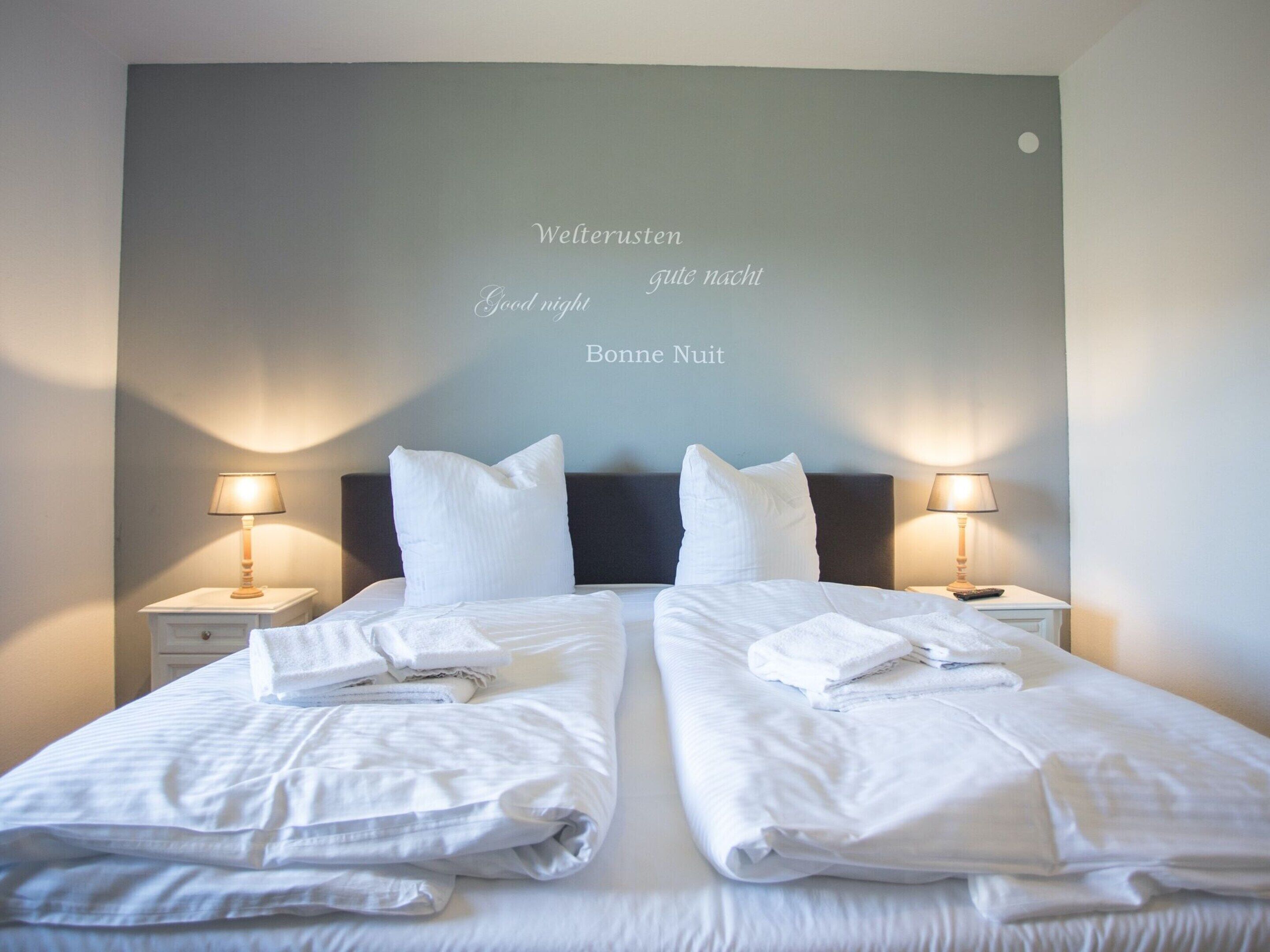 Un lit double avec du linge de lit blanc et des oreillers, deux lampes de chevet et un mur orné d’un message « Bonne nuit » dans plusieurs langues.