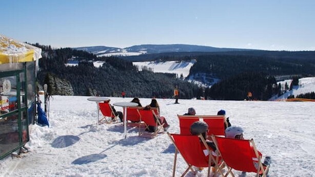 Un espace extérieur aménagé avec des sièges, recouvert de neige, avec des chaises et des tables rouges, offrant une vue sur un paysage montagneux sous un ciel dégagé.