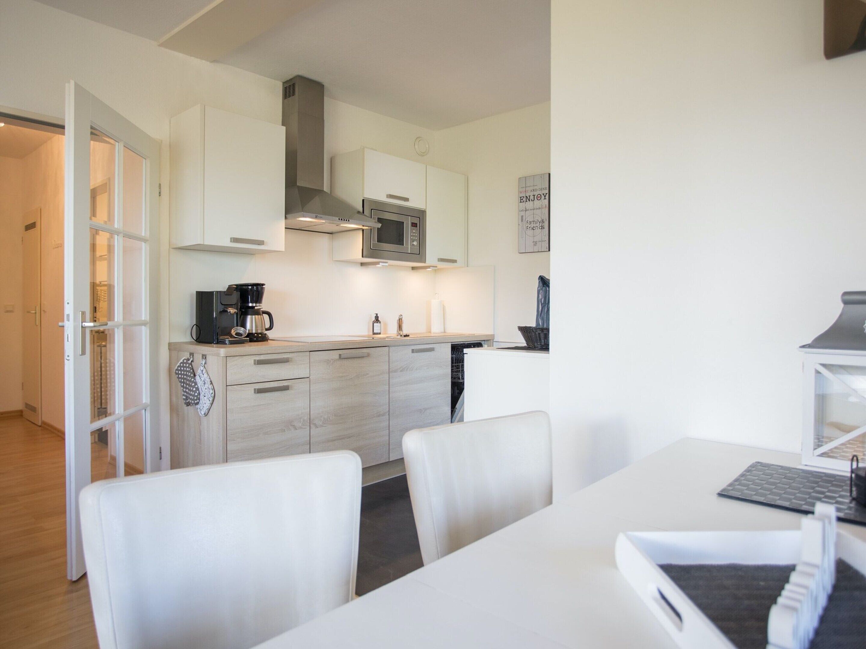 Una cocina moderna con armarios blancos, una campana extractora de acero inoxidable y un microondas. Hay una zona de comedor con una mesa y sillas blancas.