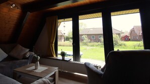 A cozy room with a sloped ceiling, a wooden table, a sofa, and a chair. A large window offers a view of a house and a barn in the distance. - Ferienhaus Amalie, Burhave (Butjadingen)