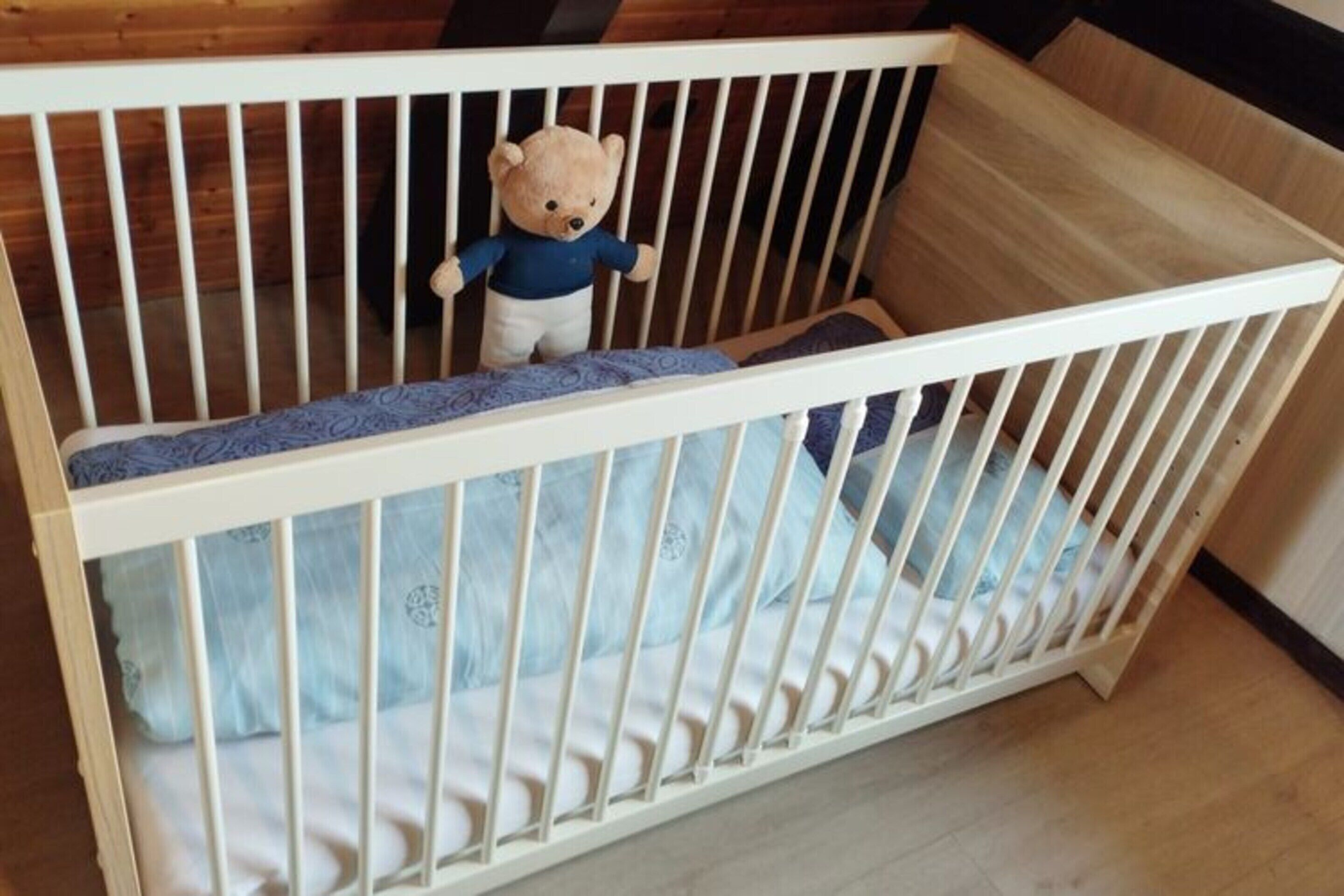 A white crib with a teddy bear inside, a blue patterned mattress, and a wooden floor.