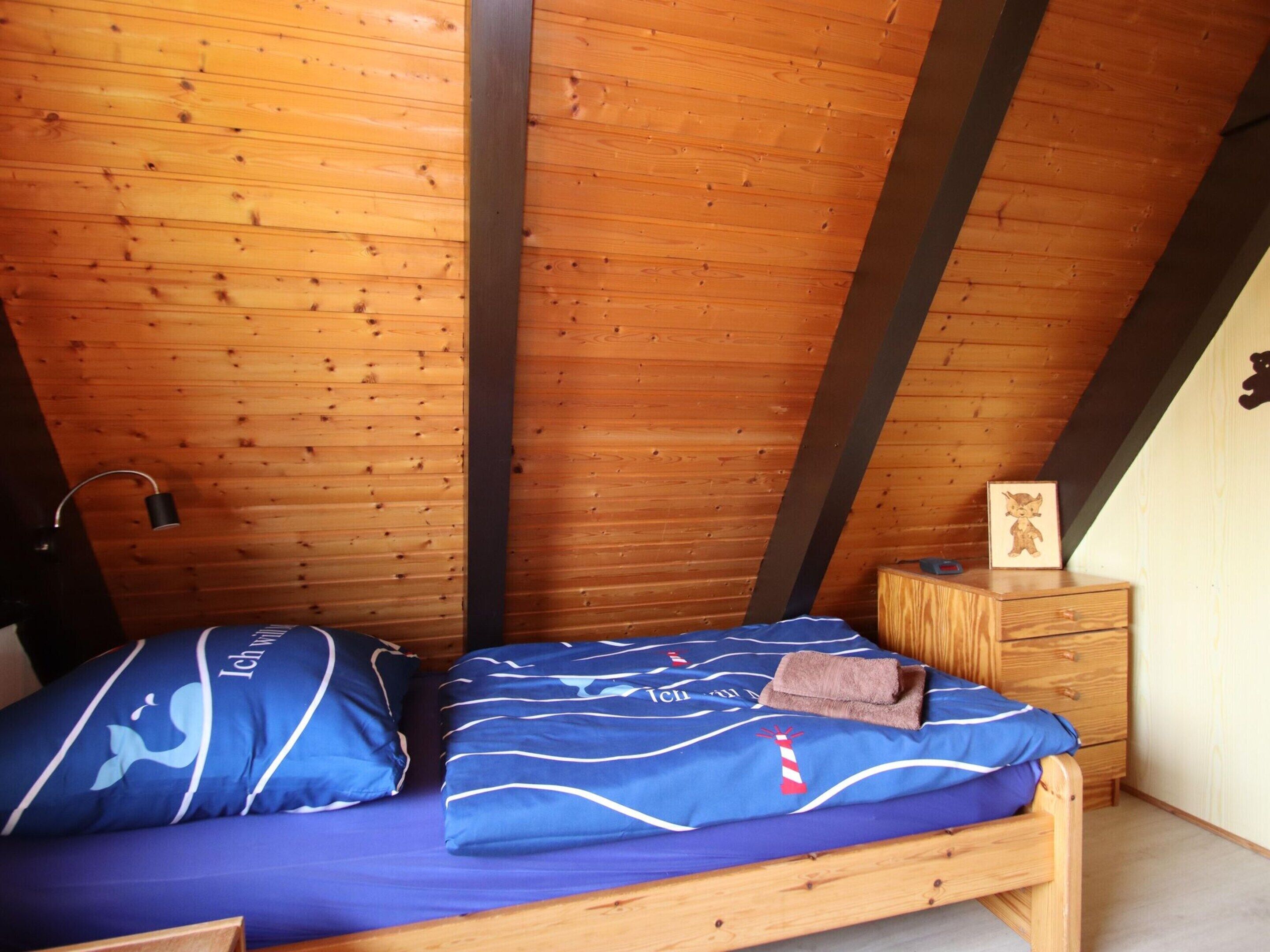 Una habitación pequeña y acogedora con techo inclinado y paredes de madera. Cuenta con una cama individual con ropa de cama azul y una mesita de noche de madera con un marco de fotos.