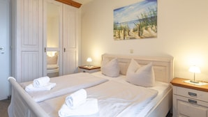 Een tweepersoonsbed met witte lakens en kussens, een houten hoofdbord, een schilderij van een strandlandschap en een nachtkastje met een lamp.