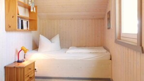 Une petite pièce agréable et chaleureuse, avec un lit simple, une table de chevet en bois, une étagère remplie de livres et de figurines, et une fenêtre laissant entrer la lumière naturelle.