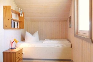 A small, cozy room with a single bed, a wooden nightstand, a shelf with books and figurines, and a window providing natural light.
