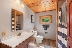 Un baño con techo de madera, un lavamanos blanco, un inodoro y una ventana con vistas a árboles.