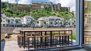 Une terrasse avec des places assises extérieures, donnant sur une ville historique aux bâtiments en pierre et sur une colline surmontée d’un château.