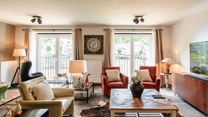 Un salon moderne comprenant un canapé, deux fauteuils, une table basse et un téléviseur. Il y a deux fenêtres avec des rideaux, une horloge au mur et une lampe sur une table d’appoint.