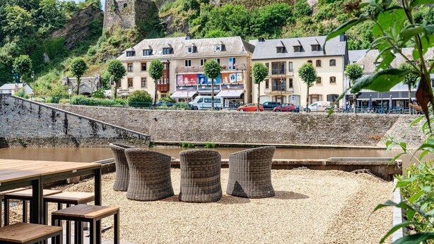 Une vue sur le fleuve, avec un château historique en arrière-plan, une rangée de bâtiments abritant des boutiques et des tables en plein air avec des chaises en osier.
