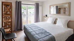Une chambre d’hôtel avec un lit, une chaise, une cloison en bois, une fenêtre avec des rideaux, un miroir et une vue sur les bâtiments environnants.