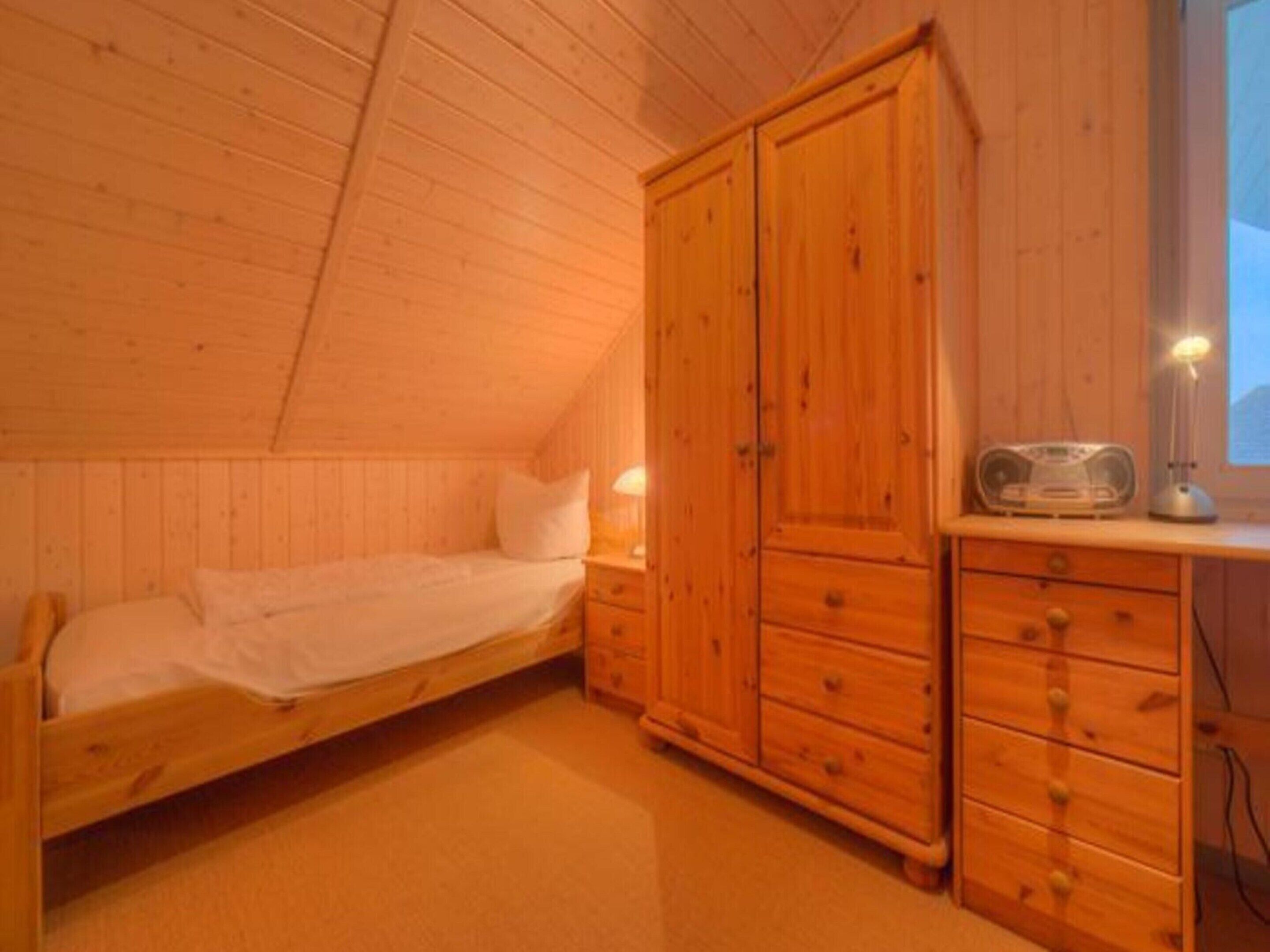 A small, cozy room with a single bed, wooden wardrobe, and a desk with a lamp.