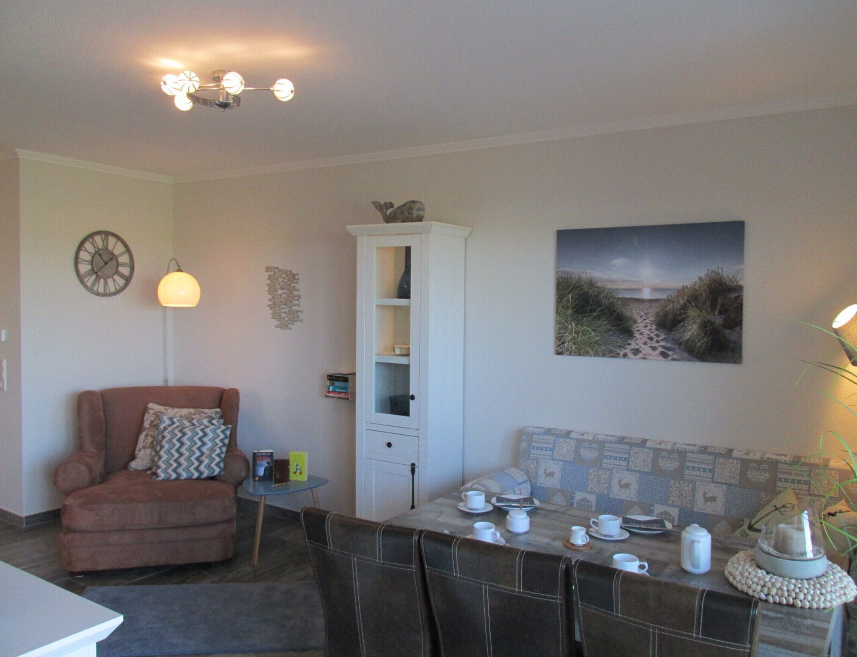 A cozy living room with a brown armchair, a dining table set with cups and saucers, a cabinet, and a wall clock.