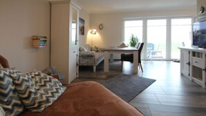 A living room with a brown sofa, a dining area with a table and chairs, and a kitchen area with cabinets and a television. - Ferienhaus mit Garten (Dornum)