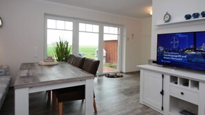 A modern kitchen with a large window, a dining area with a table and chairs, and a flat-screen TV mounted on the wall. - Ferienhaus mit Garten (Dornum)