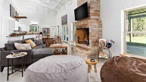Ein modernes Wohnzimmer mit Kamin aus Stein, einem Wand-Fernseher, einem grauen Sofa und einem Essbereich mit Tisch und Stühlen.
