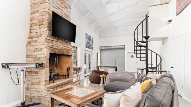 Ein modernes Wohnzimmer mit Kamin aus Stein, einem flachbildschirm-Fernseher darüber, einem grauen Sofa, einem Holztisch und einer Wendeltreppe im Hintergrund.
