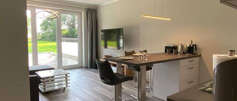A modern kitchen with a dining area, large windows, and a flat-screen TV.