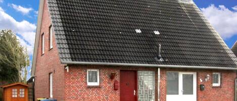 A red brick house with a steeply pitched roof, a front door, and a small front yard.