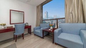 A hotel room with a desk, two armchairs, a large window with curtains, a mirror, and a vase with flowers.