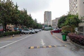 Un parking rempli de voitures, un grand immeuble en arrière-plan et une poubelle verte sur la droite.