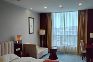 A hotel room with a bed, a sofa, a desk, and a chair. There is a window with curtains and a view of the city.