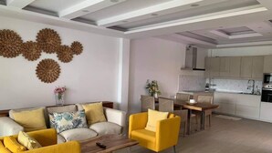 Een moderne woonkamer met een bank, fauteuils en een salontafel. Er is een eethoek met een tafel en stoelen. De keuken is op de achtergrond zichtbaar, met kasten en apparatuur.