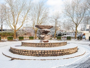 Une fontaine circulaire en pierre, recouverte de neige, à plusieurs niveaux, entourée d’un chemin circulaire pavé de briques.