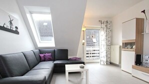A modern loft apartment with a gray sofa, a small white table, and a view of the city through a large window. - Maisonette Wohnung Meerjungfrau, Dornumersiel (Dornum)
