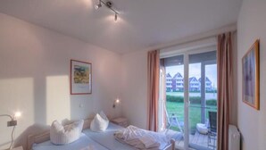A bedroom with a large bed, a bedside table, a window with curtains, and a view of buildings outside.