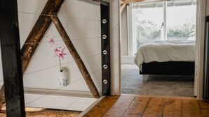 A bedroom with a bed, a wooden floor, a white wall with a decorative shelf, and a window with a view of greenery.