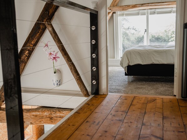 A bedroom with a bed, a wooden floor, a white wall with a decorative shelf, and a window with a view of greenery.