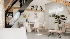 A modern loft with exposed wooden beams, a dining area with white chairs, a large potted plant, and a staircase.