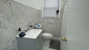 Un bagno con parete in marmo, un lavandino, un water, una finestra con persiane e un piccolo tappeto.