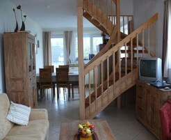 Amplia sala de estar con una escalera de madera, una mesa de comedor con sillas y un televisor sobre un mueble de madera.