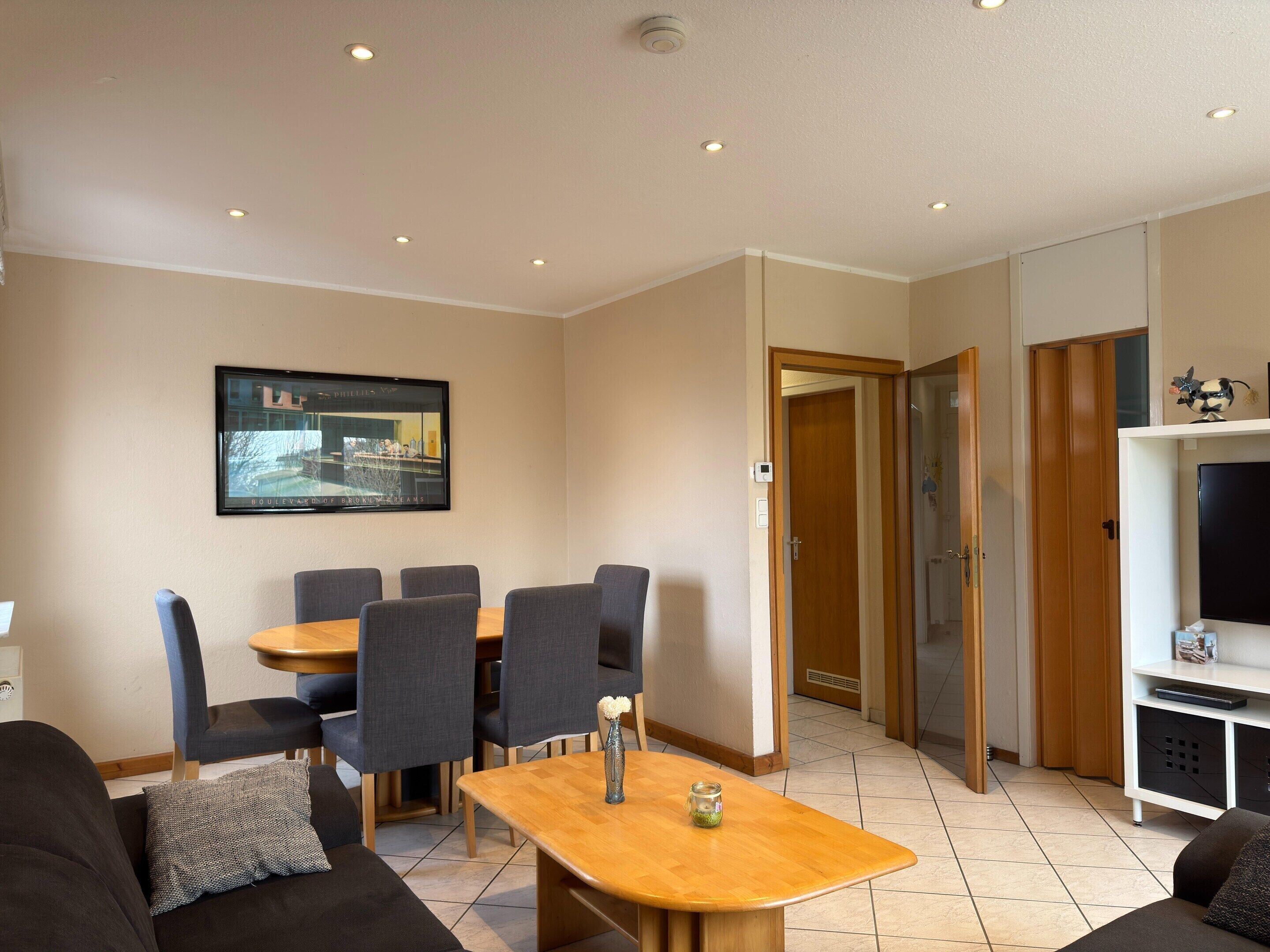 A modern living room with a dining table, chairs, sofas, a TV, and a door leading to another room.