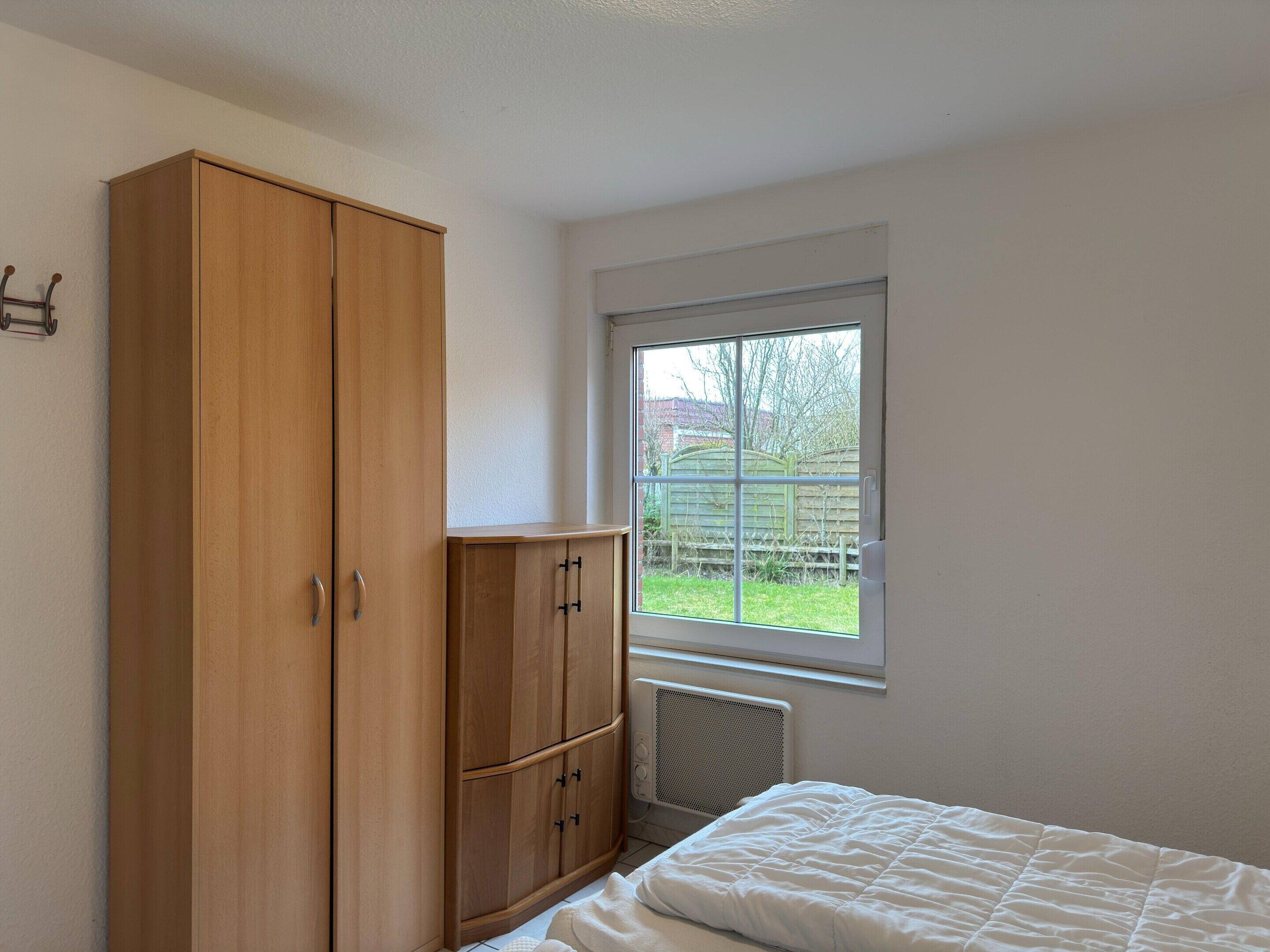 Ein Schlafzimmer mit Bett, Schrank, Heizkörper und einem Fenster mit Gartenblick.