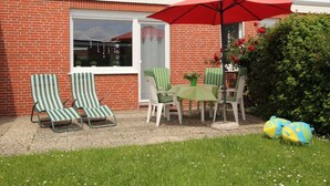 Eine Terrasse mit zwei gestreiften Liegestühlen, einem Tisch mit rotem Sonnenschirm und einem kleinen Gartenbereich mit Blumen und einem Busch.