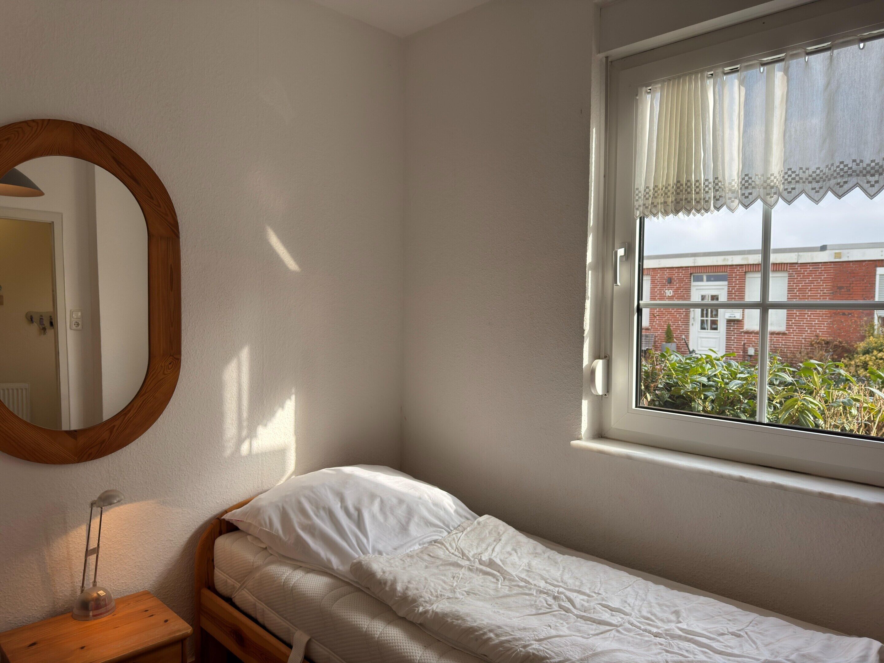 A single bed with a white mattress and pillow, a wooden bedside table with a lamp, a round mirror, and a window with a view of a brick building.